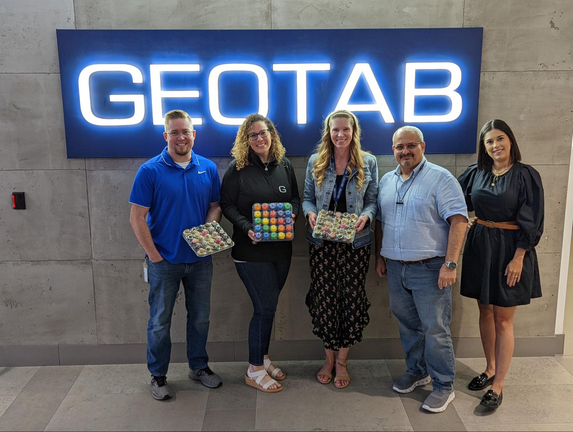 Five Geotabbers standing in front of a Geotab sign, holding Pride-themed baked goods.