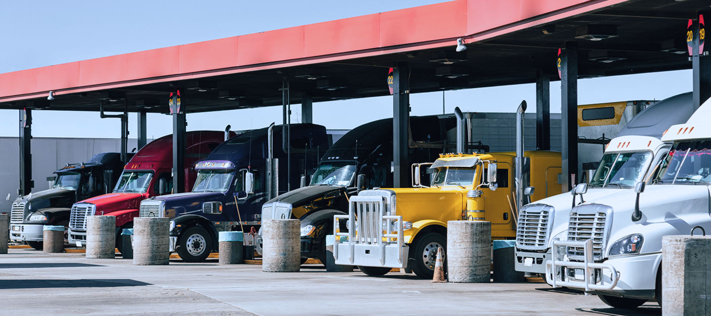 A group of fleet trucks parked at a rest stop under a red metal awning during the day, taking a break on a long-haul route.