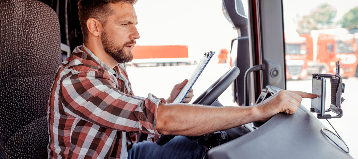 A driver in a semi-truck looking at a clip board and typing into a navigation system.