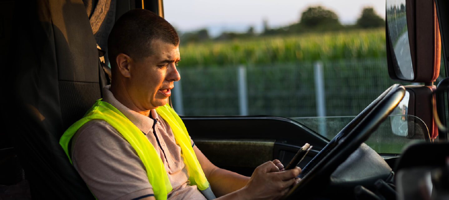  A male driver wearing a yellow safety vest behind the wheel of a fleet vehicle looking at his phone.