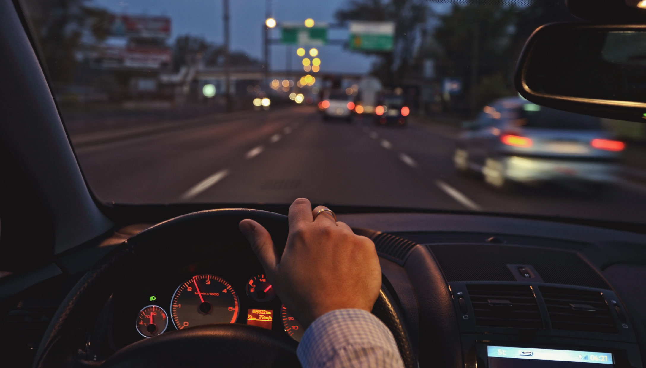 Vista desde el interior de un vehículo circulando por una avenida al anochecer, con la mano del conductor sobre el volante y varios autos al frente. La imagen representa la importancia de mantener la atención en el camino y prevenir riesgos al manejar.