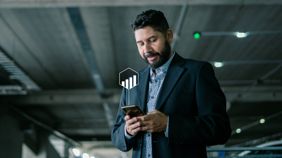 Hombre de negocios con barba usando el celular en un estacionamiento techado, vestido con saco oscuro y camisa estampada, concentrado en la pantalla de su teléfono.