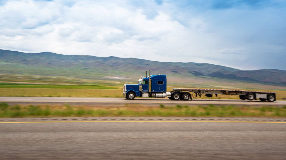 Camión de carga azul con remolque vacío en carretera montañosa bajo un cielo parcialmente nublado, representando la logística de transporte.