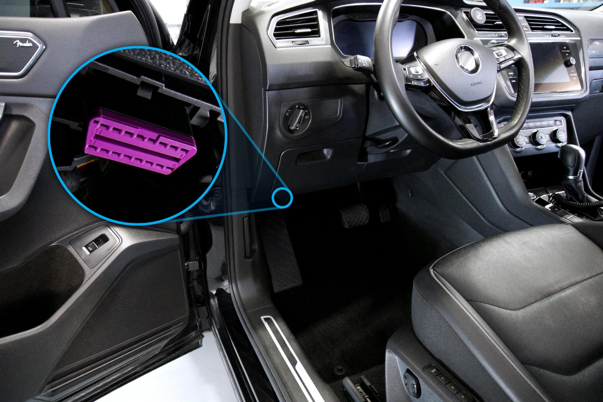 Imagen del interior de un vehículo, destacando el puerto OBD-II (On-Board Diagnostics) debajo del tablero, cerca del volante. La conexión OBD-II, resaltada en color morado, se utiliza para diagnósticos automotrices y monitoreo de datos del vehículo.