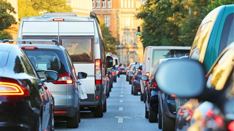 Verkehrsstau auf einer Stadtstraße, Blick auf eine Fahrspur mit Stoßstange-an-Stoßstange-Autos, darunter ein SUV und ein weißer Lieferwagen. Im Hintergrund säumen Gebäude und Bäume die Straße, ein grünes Verkehrssignal ist sichtbar. Die Beleuchtung deutet auf den späten Nachmittag hin.