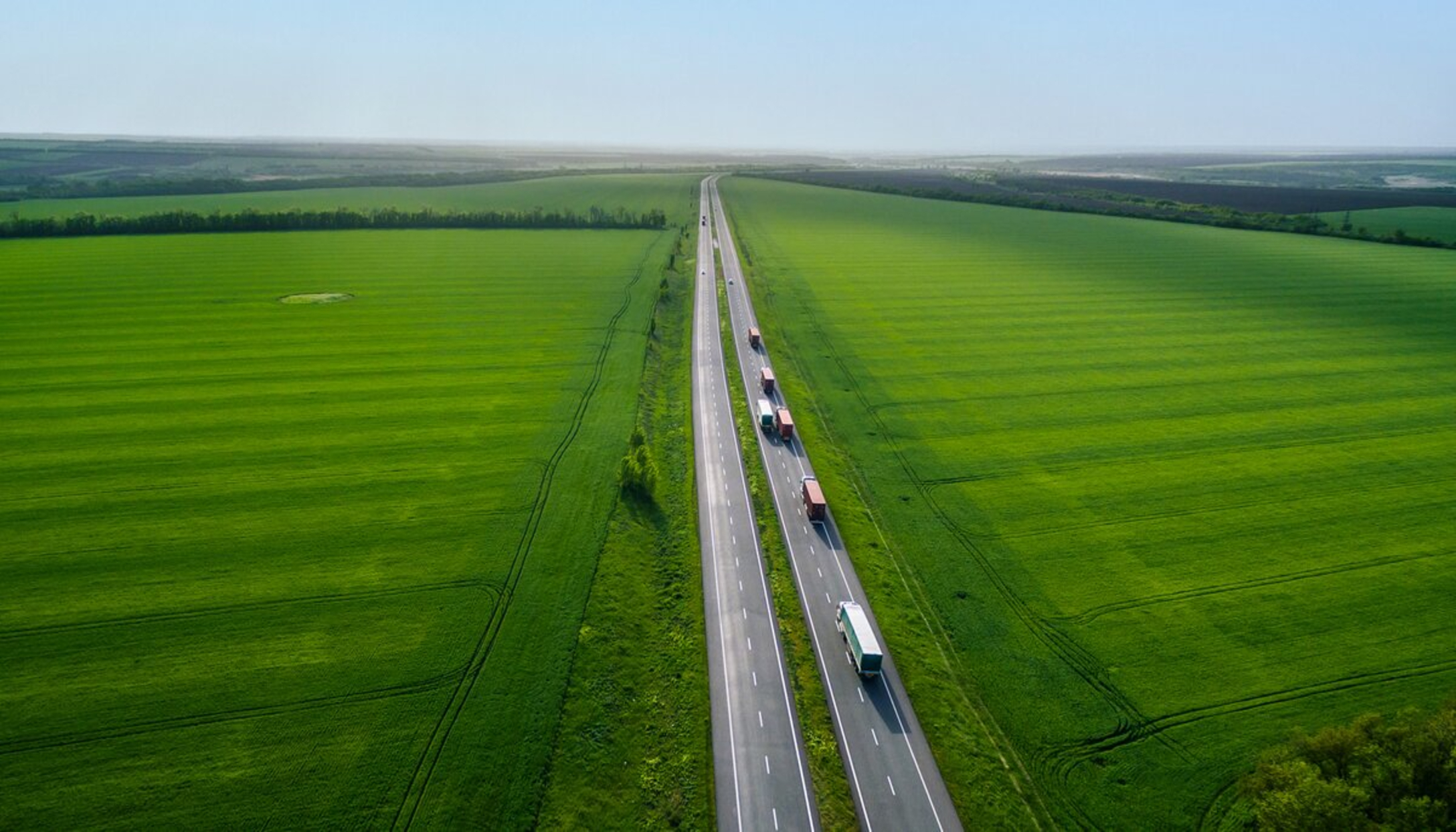 Een rij vrachtwagens rijdt over een lange, rechte weg, omringd door een uitgestrekt groen landschap.