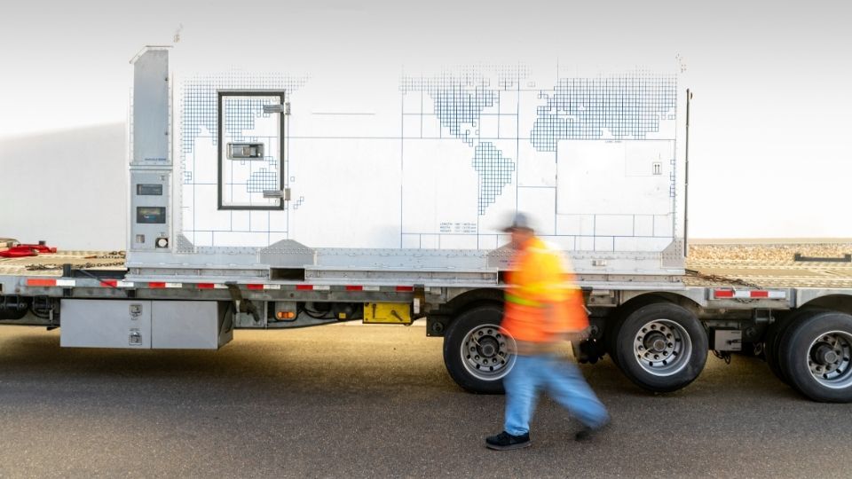This is a photograph of a large, white, rectangular container secured to a flatbed semi-trailer.